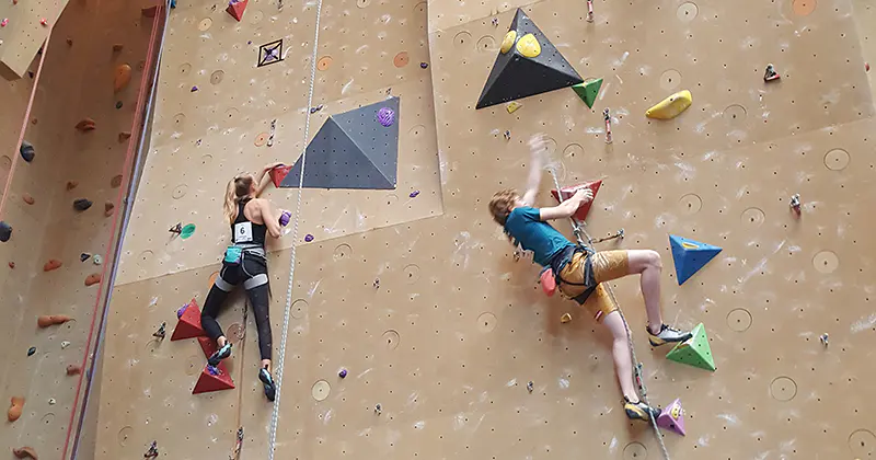 Twee mensen zijn aan het rotsklimmen op een indoor-klimwand met gekleurde grepen en geometrische vormen; ze dragen allebei een klimgordel en sportkleding.